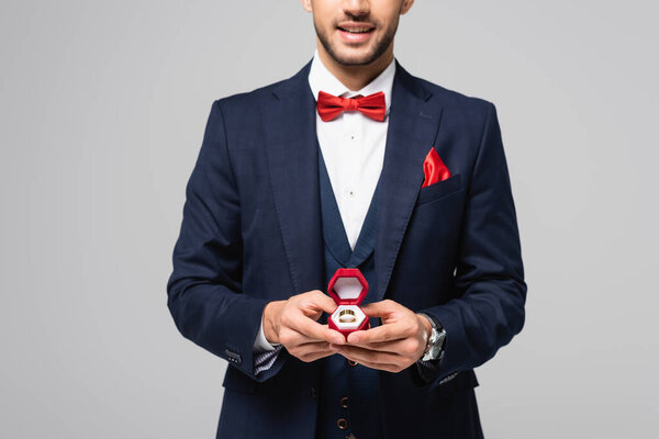 partial view of smiling man in elegant suit holding jewelry box with wedding ring isolated on grey