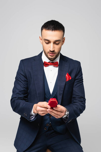 elegant hispanic man in blue suit looking at red jewelry box isolated on grey
