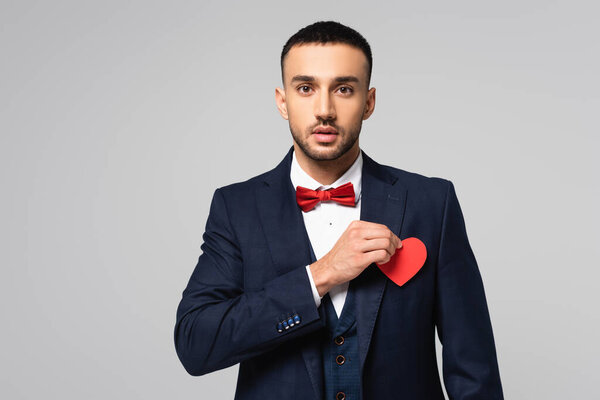 elegant hispanic man looking at camera while holding red paper cut heart isolated on grey