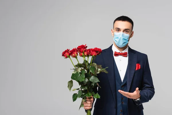 青いスーツを着た若いヒスパニック系の男とグレーに孤立した赤いバラを指している医療マスク — ストック写真