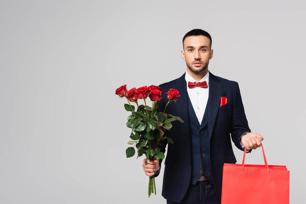 hispanic man in elegant suit looking at camera while holding red roses and shopping bags isolated on grey