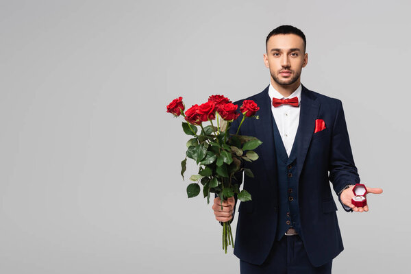 elegant hispanic man in blue suit holding red roses and jewelry box isolated on grey