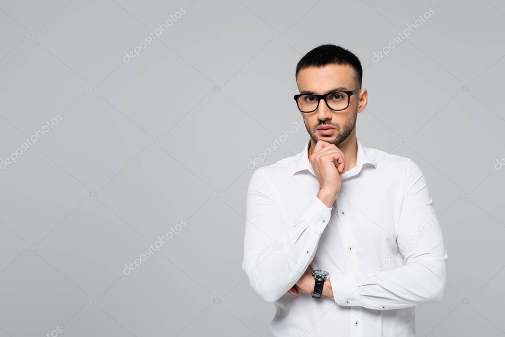 thoughtful hispanic manager holding hand near chin while looking at camera isolated on grey
