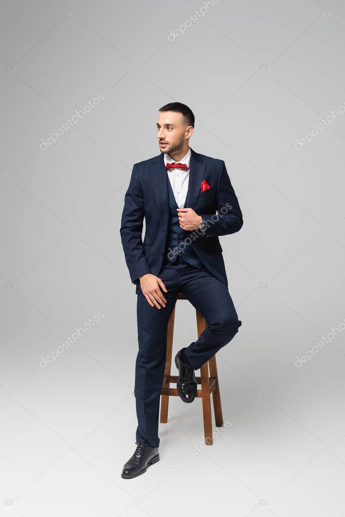 full length view of young hispanic man in elegant suit posing on high stool on grey