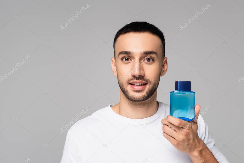 brunette hispanic man holding vial of eau de cologne and looking at camera isolated on grey