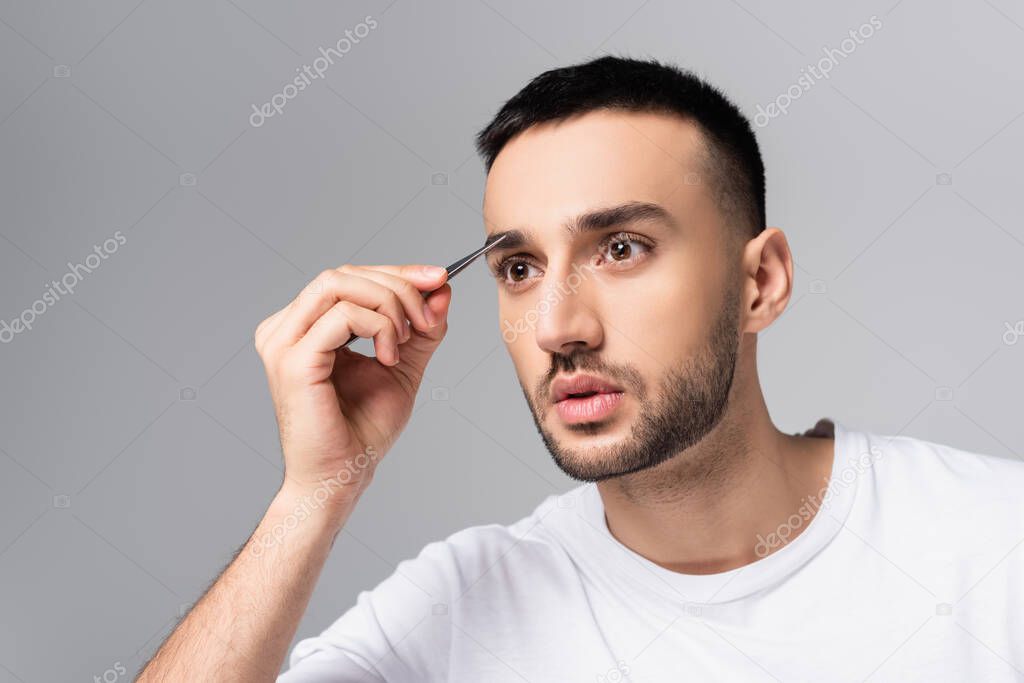 young hispanic man tweezing eyebrows isolated on grey