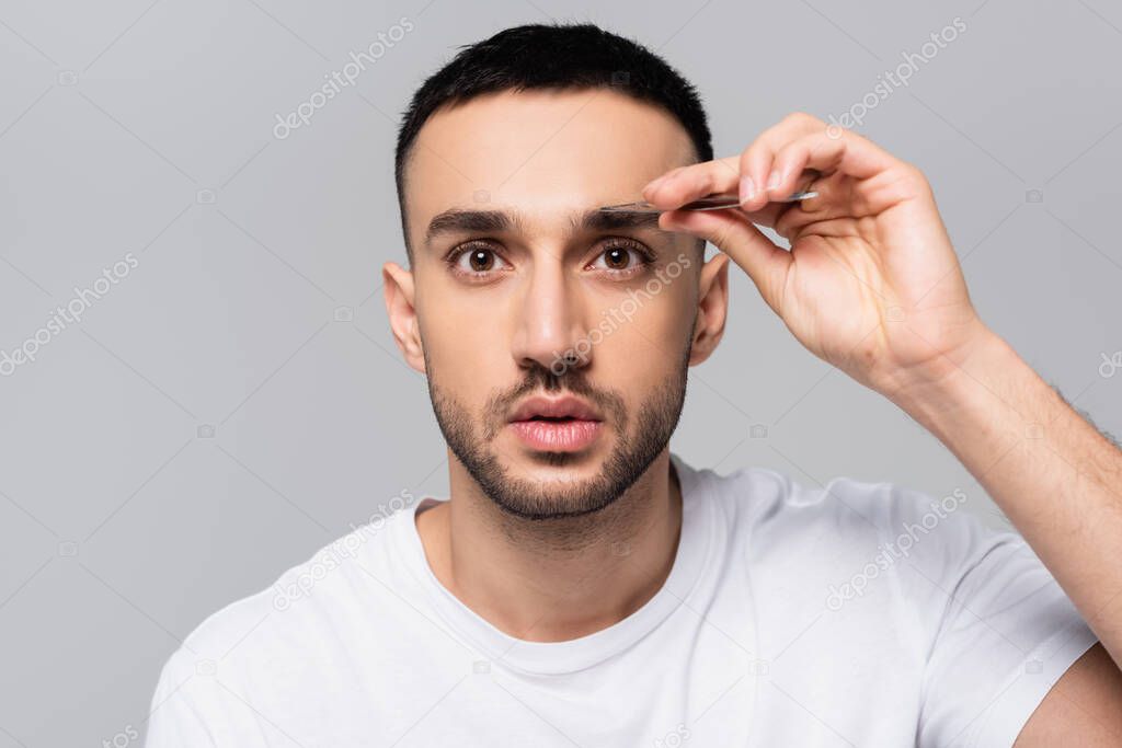 brunette hispanic man looking at camera while tweezing eyebrows isolated on grey