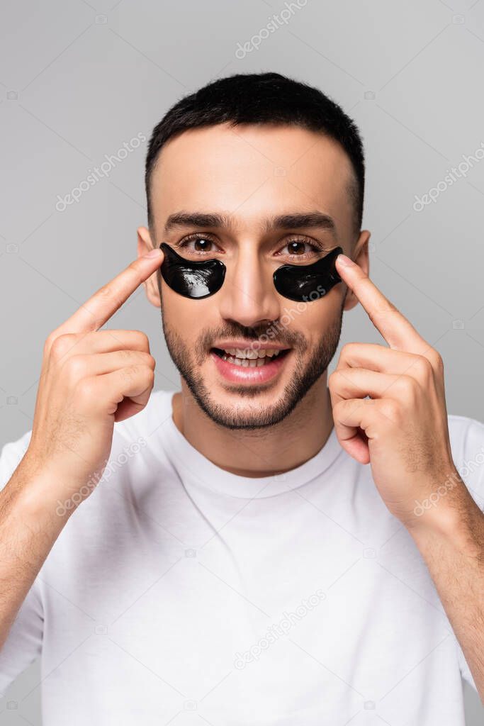 smiling hispanic man applying eye patches isolated on grey