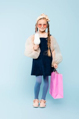 Güneş gözlüklü, elinde bardak, elinde milkshake ve mavi çantalı küçük bir kız çocuğu.