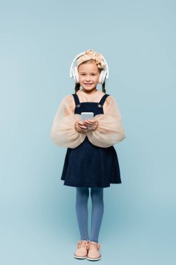 Mavi renkte izole akıllı telefon kullanan, yaylı ve kablosuz kulaklıklı neşeli çocukla dolu. 