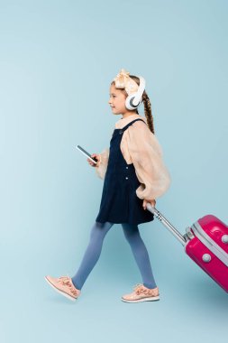 Kablosuz kulaklıklı küçük bir kız elinde akıllı bir telefonla mavi bavulla yürürken. 