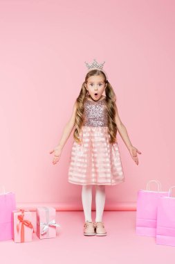 full length of shocked little girl in crown standing near presents and shopping bags on pink  clipart