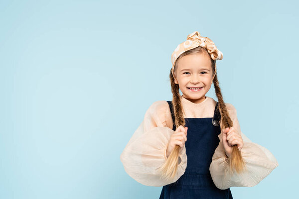 cheerful kid touching pigtails and smiling isolated on blue