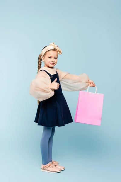 幸せな子供の完全な長さピグテールショッピングバッグを保持し 青い上に親指を表示 — ストック写真