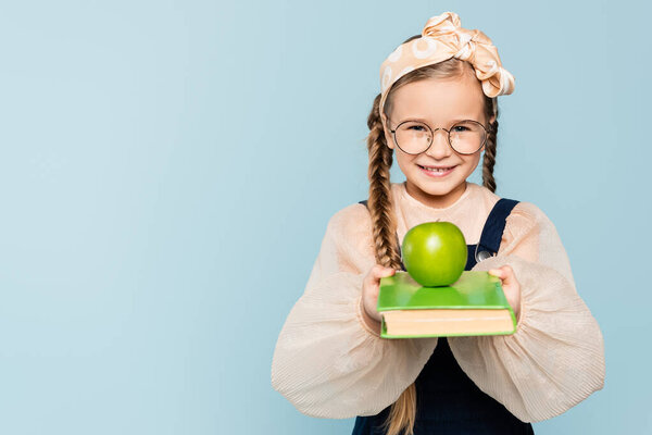 smart kid in glasses holding book with green apple and smiling isolated on blue