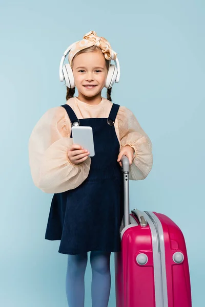 Szczęśliwa Dziewczynka Słuchawkach Bezprzewodowych Pomocą Smartfona Podczas Stania Bagażem Niebiesko — Zdjęcie stockowe