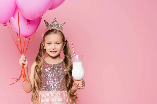 穿着衣服 头戴皇冠的快乐小女孩拿着杯子 奶昔和气球与粉色隔离在一起 — 图库照片