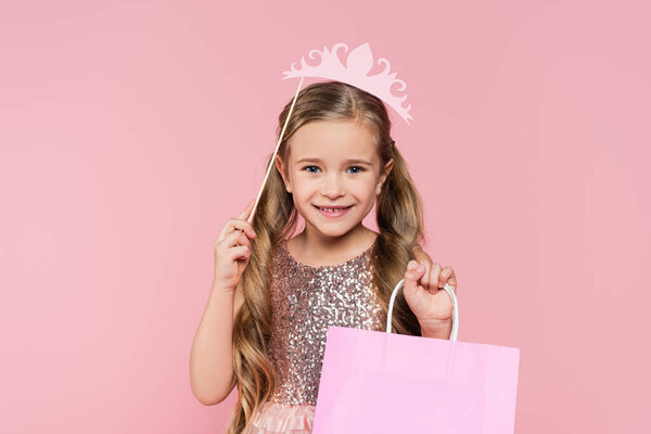 cheerful little girl in dress holding paper crown on stick above head and shopping bag isolated on pink 