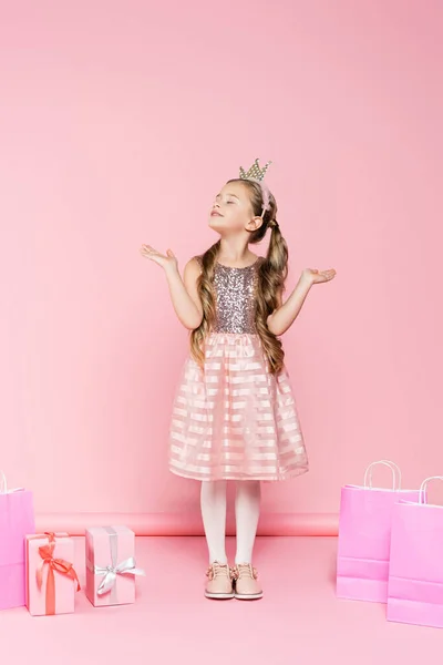 満足している小さな女の子の王冠とともに閉じられた目の近くに立ってプレゼントやショッピングバッグオンピンク — ストック写真