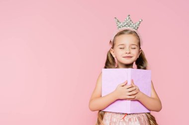 smiling little girl with closed eyes in crown holding book isolated on pink clipart