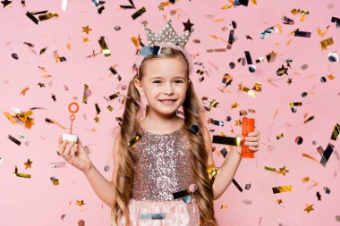 Taçlı neşeli küçük kız pembe üzerine düşen konfetilerin yanında sabun köpükleri tutuyor. 