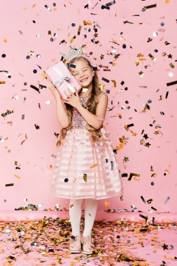 full length of happy little girl in crown holding present near falling confetti on pink  clipart