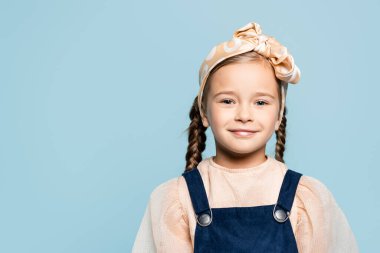 cheerful kid in headband with bow looking at camera isolated on blue clipart