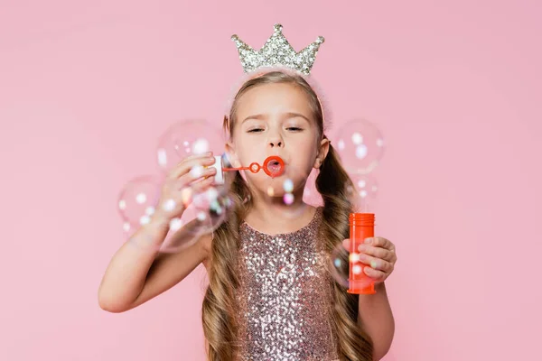 戴着皇冠的小女孩在模糊的前景上吹着肥皂泡 与粉色隔离 — 图库照片