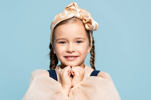 positive kid in headband with bow looking at camera isolated on blue