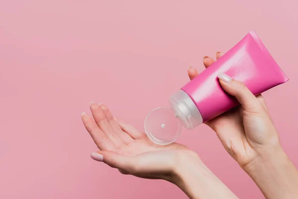 cropped view of woman holding tube while applying lotion on hand isolated on pink