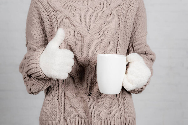 Cropped view of young woman in knitted sweater and warm gloves holding cup and showing thumb up on white background