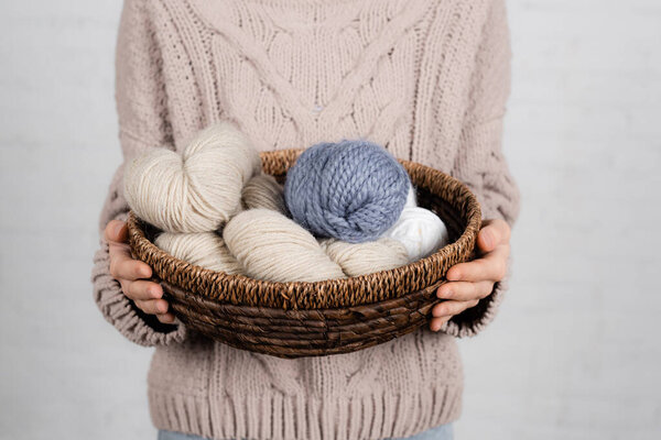 Cropped view of woman in sweater holding basket with yarn balls on white background