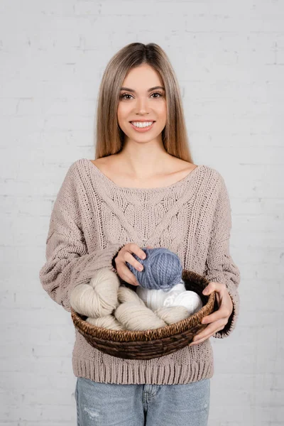 Cheerful woman in cozy sweater holding basket with woolen yarn on white background