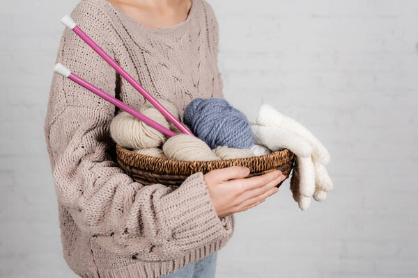 Cropped view of young woman holding basket with knitting needles, yarn and gloves on white background