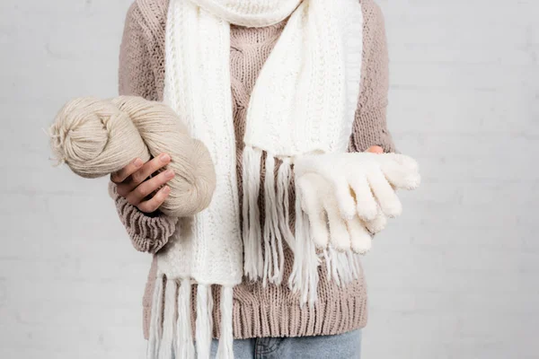 Cropped view of woman in sweater and scarf holding yarn and gloves on white background