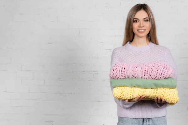 Mujer Joven Sonriendo Cámara Mientras Sostiene Suéteres Colores Con Pared — Foto de Stock