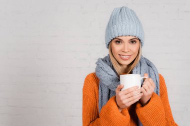Stylish woman in sweater and hat holding cup and smiling on white background clipart