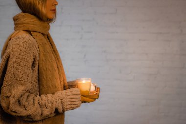 Cropped view of woman in sweater, scarf and gloves holding burning candles near white brick wall  clipart