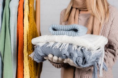Cropped view of woman holding knitwear near hanger rack with sweaters on white background clipart