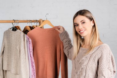 Gülümseyen kadın, beyaz arka planda askıdaki süveterlerin yanında kameraya bakıyor.