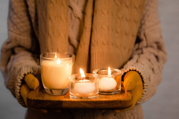 Close up view of burning candles in hands of woman in knitwear and gloves blurred on grey background