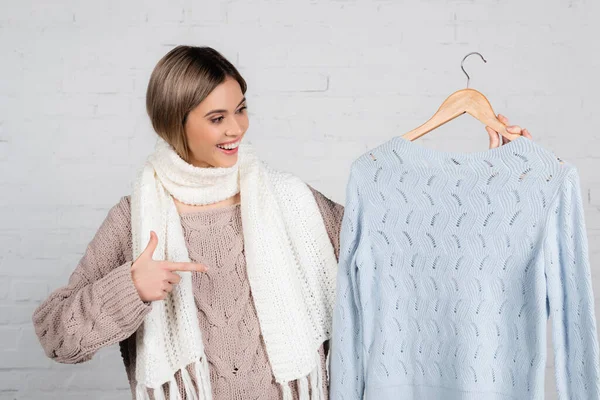 Cheerful woman in scarf pointing at hanger with sweater on white background