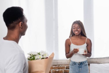 Mutlu Afro-Amerikalı kadın elinde kahve fincanı tutuyordu. Erkek arkadaşının yanında, evinin önündeki bulanık ön planda çiçekler vardı. 