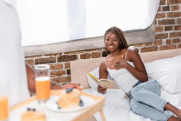 笑顔アフリカ系アメリカ人女性とともに本見ていますボーイフレンドとともに朝食でぼやけたフォアグラウンドでベッドルーム — ストック写真