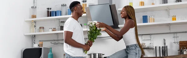 Smiling african american man presenting flowers to girlfriend in kitchen, banner