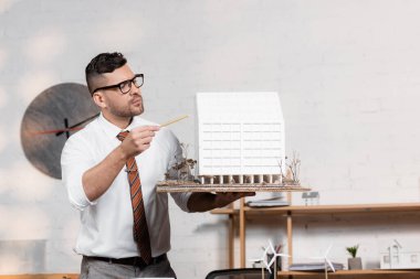 Gözlüklü ciddi bir mimar elinde kalemle ev modelini işaret ediyor.