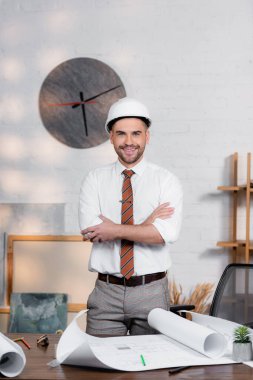 happy architect in helmet standing with crossed arms near blueprints on desk clipart