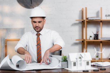 Miğferli ciddi bir mimar ev modelinin yanındaki planlara bakıyor.