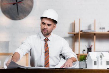 Miğferde konsantre bir mimar ev modelinin yanındaki proje üzerinde çalışıyor.