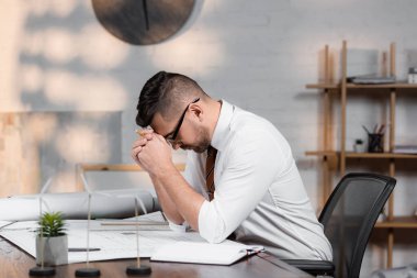 tired architect sitting with bowed head near blueprints on desk clipart